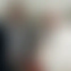 Un hombre y una figura eclesiástica conversan sonrientes en un salón amplio con techo de patrón luminoso. Ambiente cordial y formal.