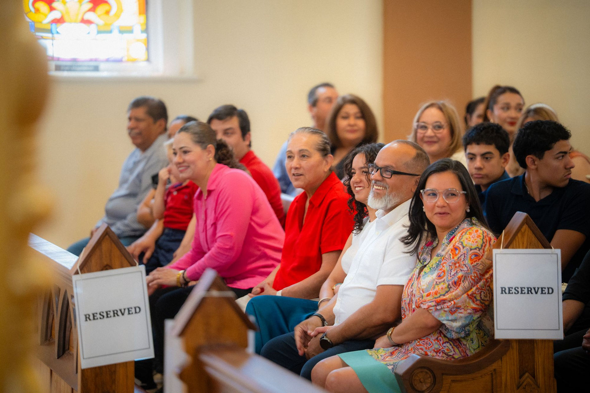 Seated congregation in a church, smiling and chatting near wooden pews with RESERVED signs.