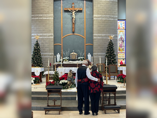 Pareja de espaldas abrazada en iglesia decorada con árboles de Navidad. Altar al fondo con crucifijo. Ambiente solemne y festivo.
