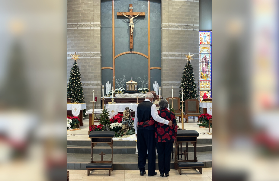Pareja de espaldas abrazada en iglesia decorada con árboles de Navidad. Altar al fondo con crucifijo. Ambiente solemne y festivo.