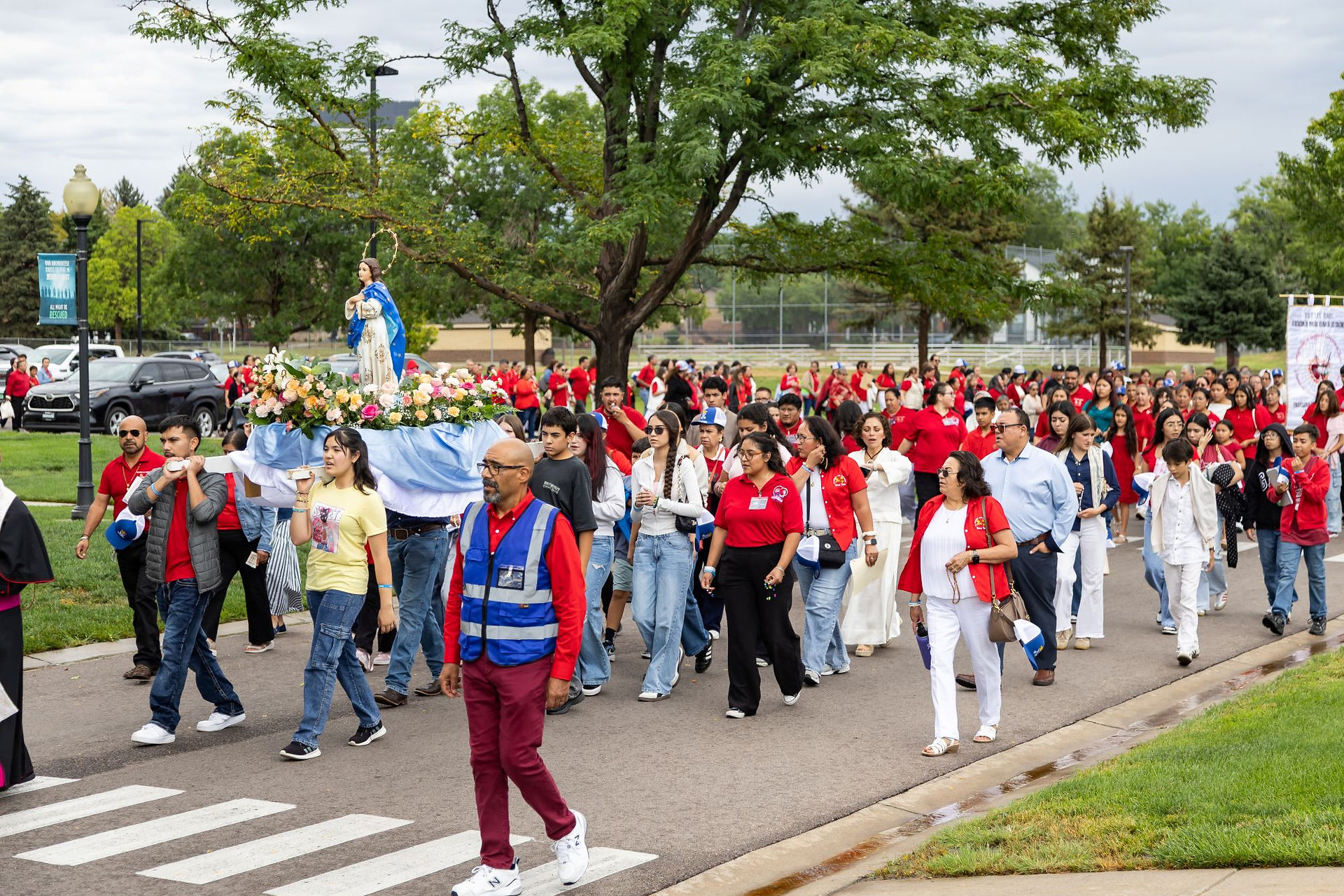 Crowd in red shirts processes through a park carrying a flower-covered statue of Mary on a platform.