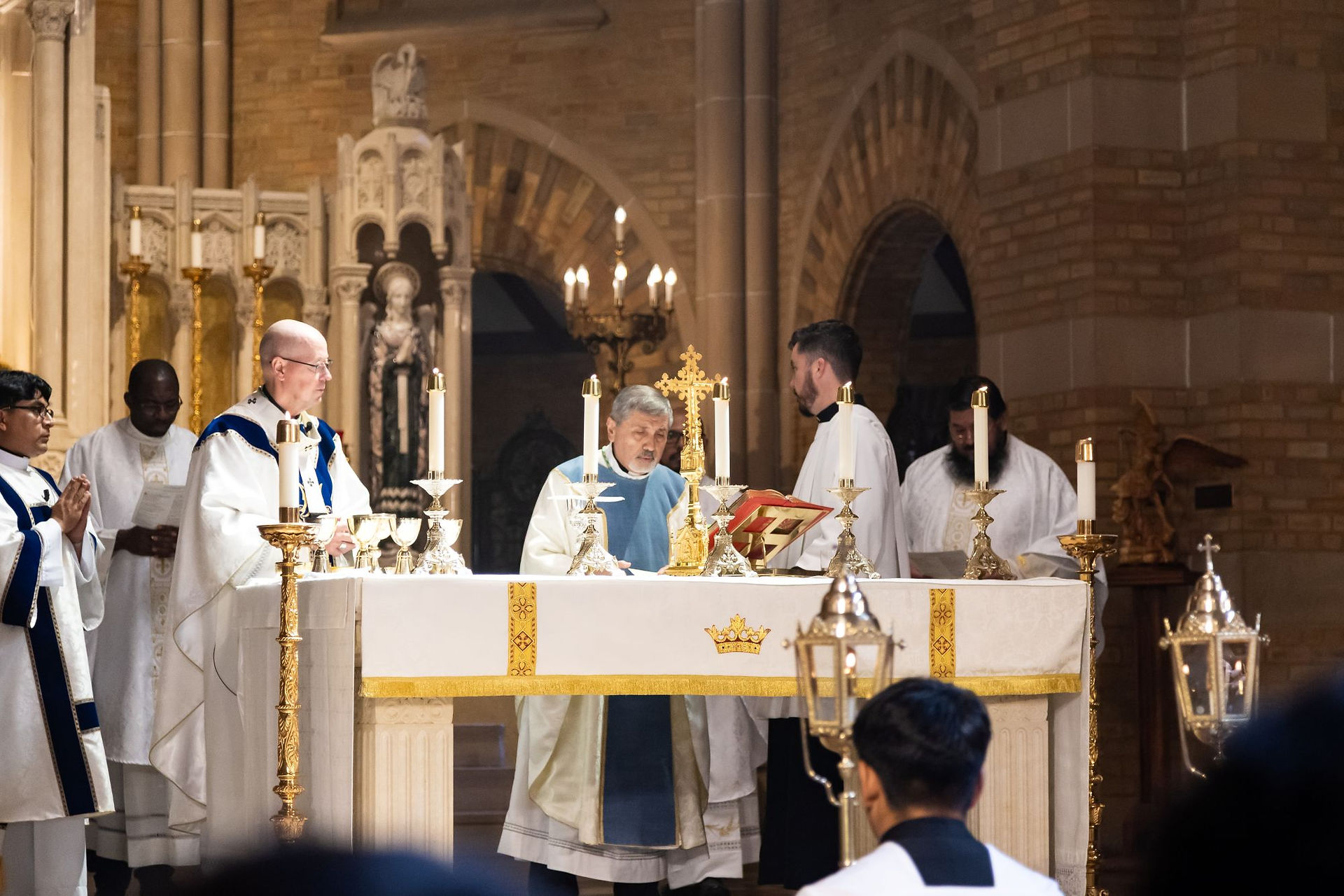 Catholic priests in white and blue vestments celebrate Mass at a candlelit altar inside a brick church.