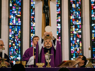 Sacerdote en misa alzando la hostia. Viste ropaje púrpura. Fondo de vidrieras coloridas. Emoción solemne.