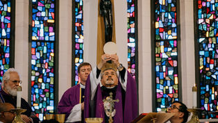 Sacerdote en misa alzando la hostia. Viste ropaje púrpura. Fondo de vidrieras coloridas. Emoción solemne.