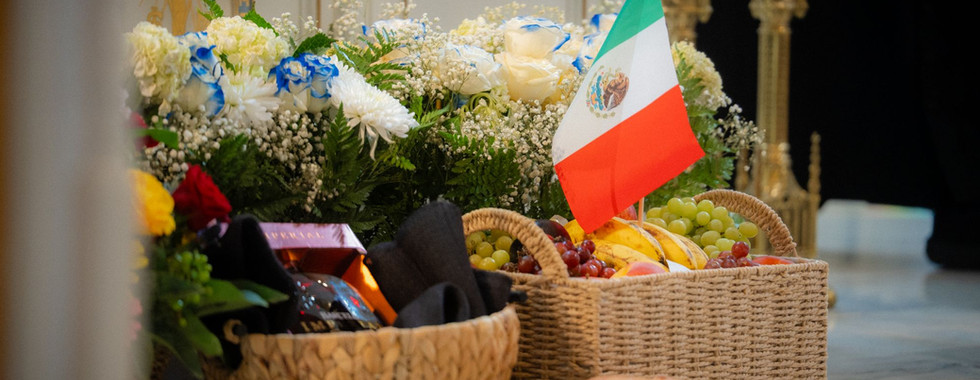Fruit baskets, bread, and white-blue flowers with a small Mexican flag in a church, suggesting a solemn tribute.