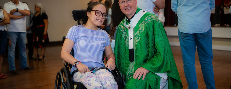 Smiling woman in a wheelchair poses with a bishop in green robes at an indoor gathering, with people standing in back.