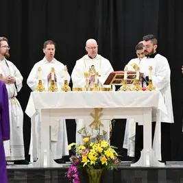 Sacerdote da la comunión a una estudiante con toga morada en un gimnasio escolar, bajo pancartas de campeonatos y un ambiente solemne.
