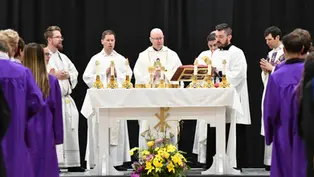 Sacerdote da la comunión a una estudiante con toga morada en un gimnasio escolar, bajo pancartas de campeonatos y un ambiente solemne.