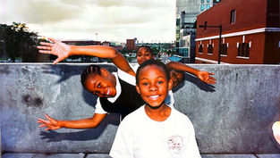 Niños sonrientes posando alegremente en una terraza urbana; edificio rojo al fondo. Ropa blanca destaca sobre el muro gris. Cielo nublado.