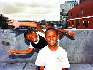 Niños sonrientes posando alegremente en una terraza urbana; edificio rojo al fondo. Ropa blanca destaca sobre el muro gris. Cielo nublado.