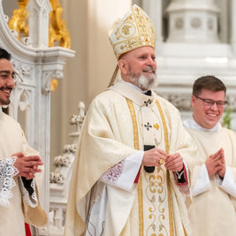 Tres clérigos sonríen en una iglesia decorada. Visten hábitos blancos y dorados, con detalles en encaje. Ambiente solemne y festivo.