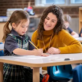 Niña y mujer en aula, la niña escribe mientras la mujer en suéter amarillo sonríe. Fondo desenfocado, ambiente educativo y cálido.