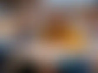Niña y mujer en aula, la niña escribe mientras la mujer en suéter amarillo sonríe. Fondo desenfocado, ambiente educativo y cálido.