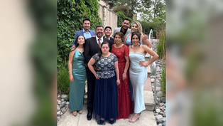 A group of nine people in formal attire pose happily outdoors, surrounded by greenery. The mood is joyful and celebratory.