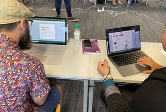 This image shows a man and a woman sitting at a table looking at computers. They are both looking at a laptop and reading each others books.