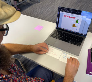 This is an image of a man holding 2 index cards that he was given for this assignment. One index card says "problem, character is really hungry" and the other card says "setting, Centra, Tesco or another Irish grocery store." He is sitting in front of a laptop and brainstorming story ideas.