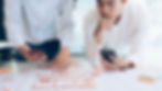 Two designers collaboratively review wireframes on a table, one pointing to a layout while the other holds a smartphone, showcasing a user-centric design process.