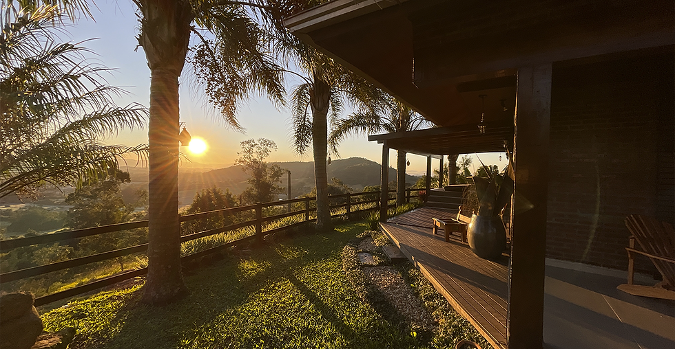 Varanda da casa refúgio ao amanhecer, com vasos decorativos e cadeiras de madeira, cercada por palmeiras e vista para montanhas ao fundo com o sol dourado nascendo