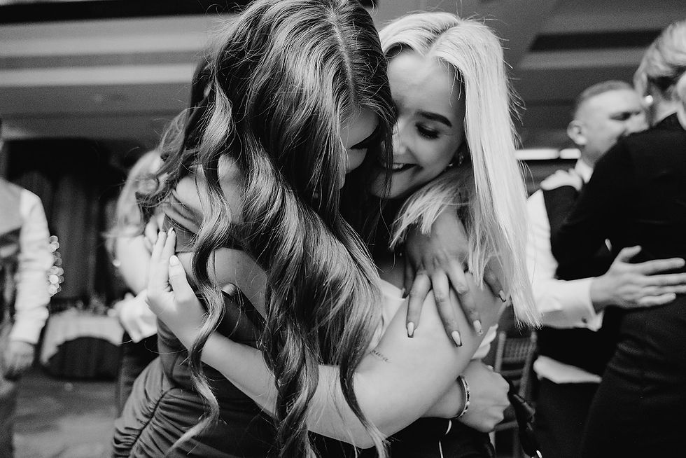Two women embrace closely, smiling in a joyful, candid moment at a social event. Black and white setting, festive background.