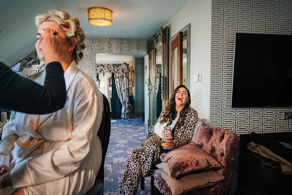 Two women in a stylish room: one getting makeup done, and the other in a leopard print outfit laughing with a drink. Warm lighting.