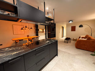 Cuisine épuipée noire avec plan de travail marbré noir et blanc, carrelage gris/beige clair avec vue sur le salon et fauteuil ocre