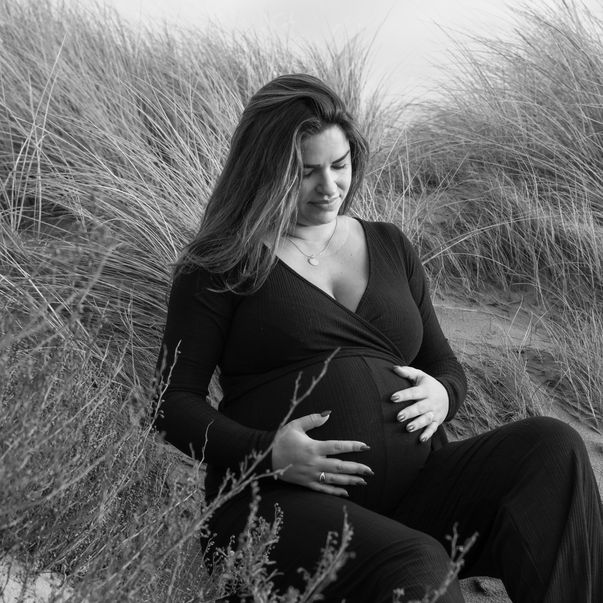 Een zwart-wit foto met een zwangere vrouw die tussen helmgras in de duinen zit. 