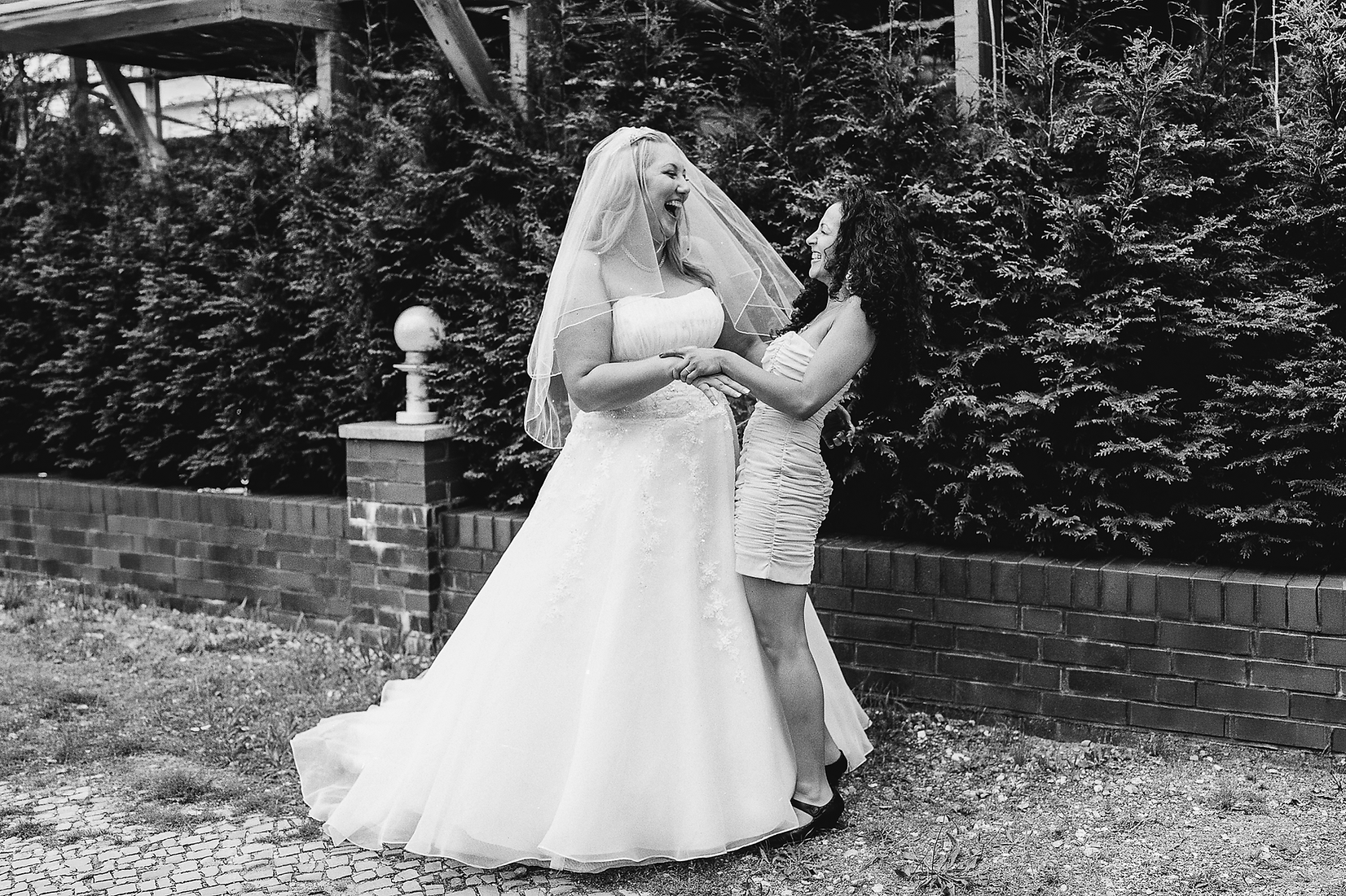 Braut im Hochzeitskleid mit Schleier lacht mit Frau im kurzen Kleid vor Hecke und Backsteinmauer.