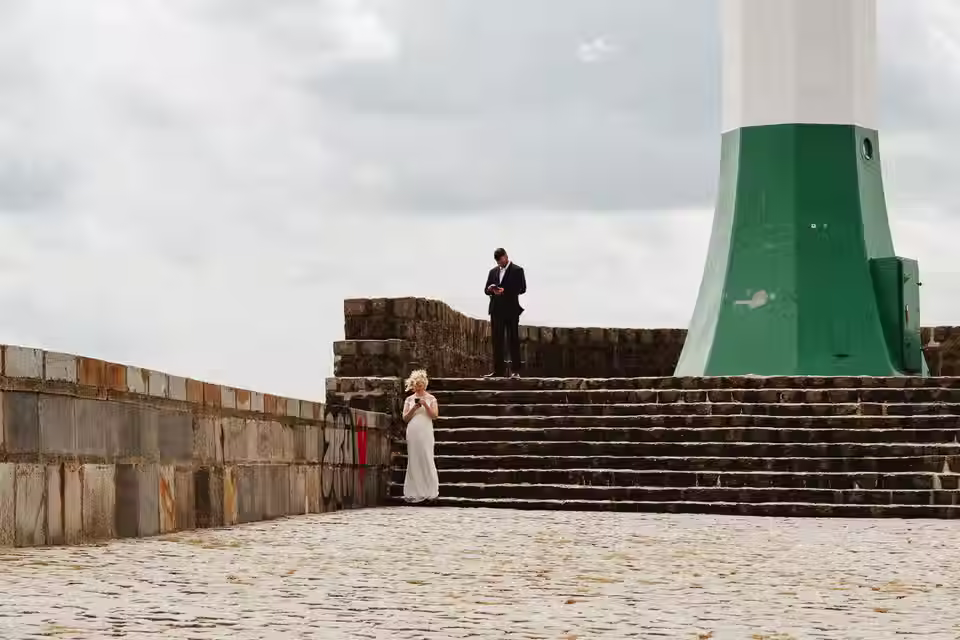 Ein Brautpaar steht mit seinen Handys in der Hand an und auf einer Treppe. Es ist der Leuchtturm von Sassnitz zu sehen.