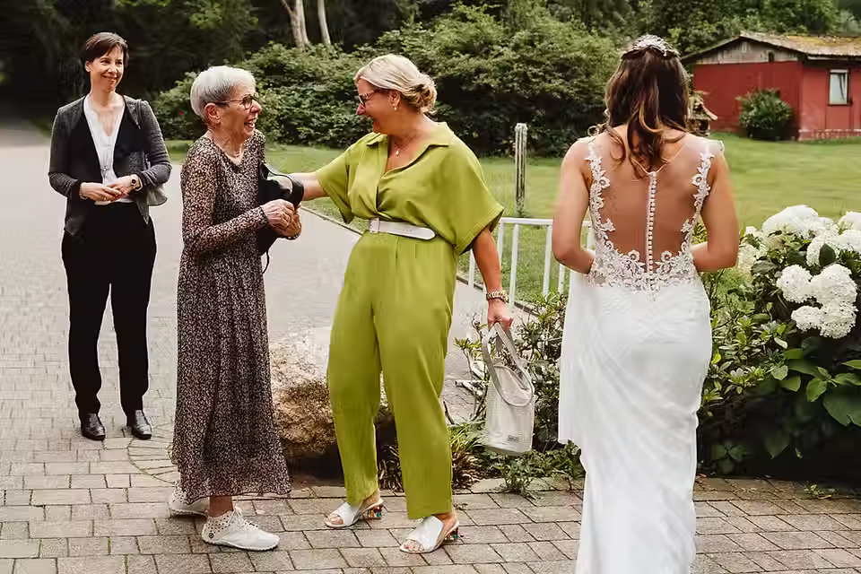 Drei Frauen begrüßen sich lachend auf einem gepflasterten Weg, eine trägt ein weißes Brautkleid mit transparentem Rückenteil und Spitzenapplikationen.