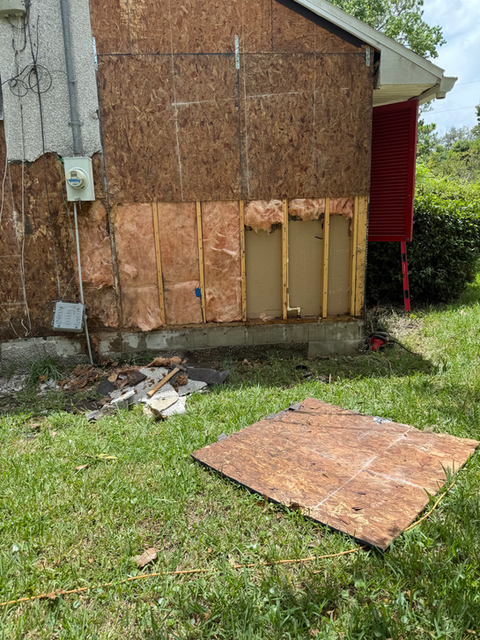 Damaged house exterior with exposed wall sections, grass, debris, and insulation.