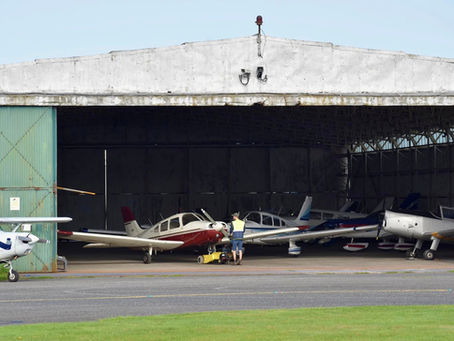 Small planes parked in a hangar, with a worker in a safety vest standing by a red-and-white aircraft on the tarmac.