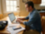 Man in denim shirt working on a laptop at a wooden table, reviewing papers and passport. Natural light from a window. Concentrated mood.