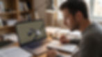 Man studies at a desk, taking notes beside a laptop playing an engineering video in a warm home office.