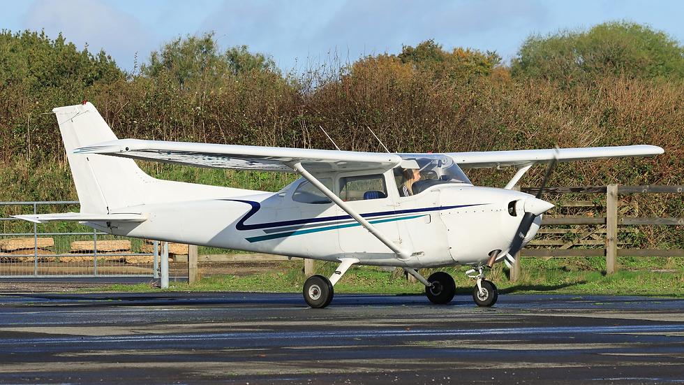 White single-engine plane with a pilot sits on a wet runway beside trees under a clear blue sky.