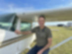 Man sitting on a small plane's wing, smiling, wearing a stethoscope. Clear sky, grassy airfield, hangars in background. Casual mood.