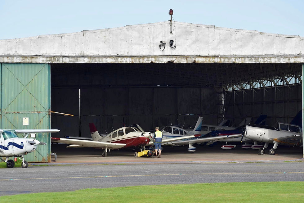 Small planes parked in a hangar, with a worker in a safety vest standing by a red-and-white aircraft on the tarmac.