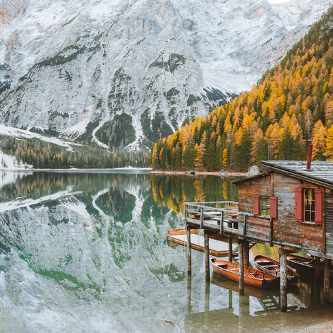 A serene view of a wooden cabin and dock on the edge of a crystal-clear alpine lake, surrounded by autumn-colored trees and snow-dusted mountains in South Tyrol.