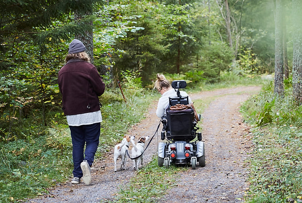 A lady in a motorised wheelchair walking her dog with her disability support carer.