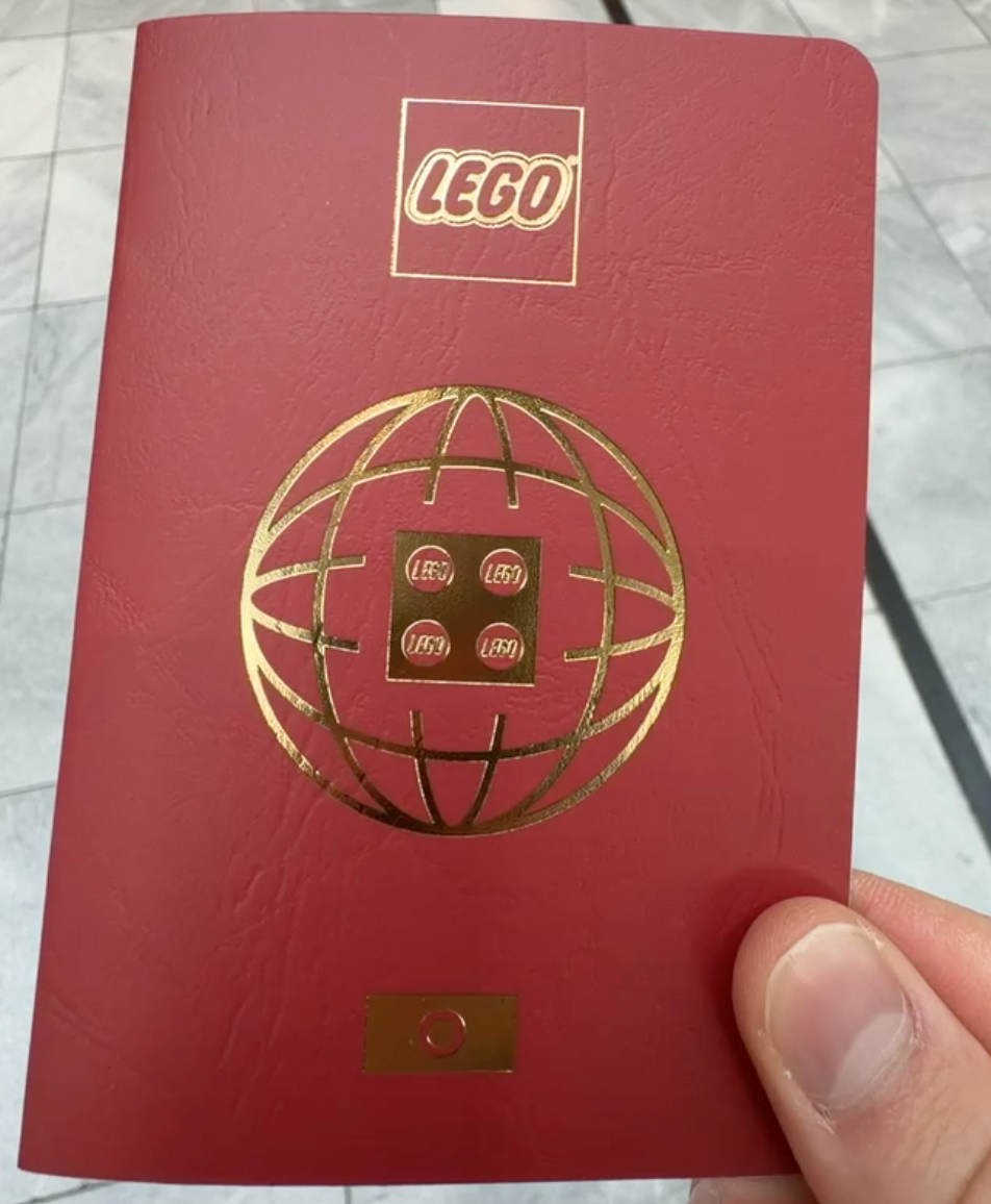 Hand holding a red LEGO Passport with a gold foil LEGO logo, globe-and-brick emblem, and small gold brick detail at the bottom, photographed outdoors on a marble tile surface.