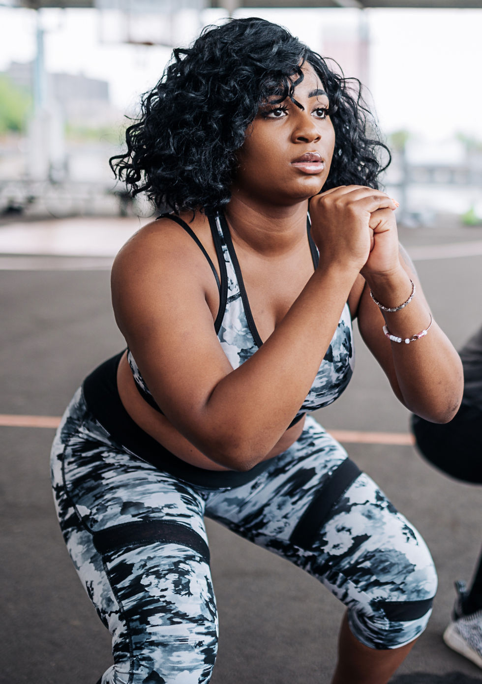 Woman squatting in a gym, wearing a black and white patterned workout outfit, focused expression. Blurred background of exercise equipment.