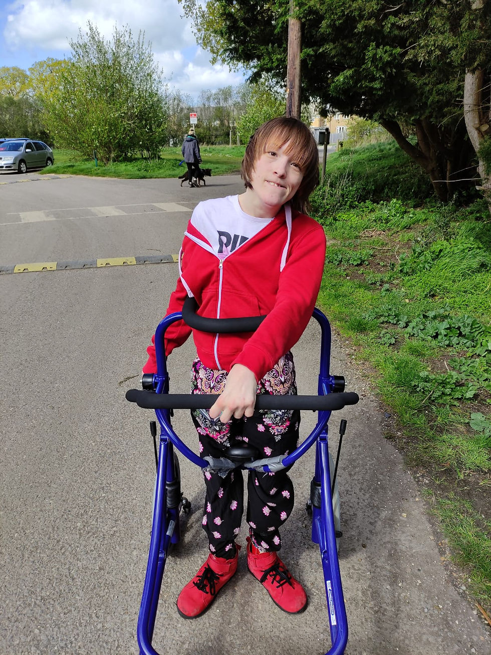 Ailsa standing in her blue walking frame. The frame has black foam around the thoracic ring and the handlebar. Ailsa is wearing a white t-shirt with Pink, the artist, on the front and wears a red hoodie with a white zip. Ailsa has black floral trousers and red boots with black laces.