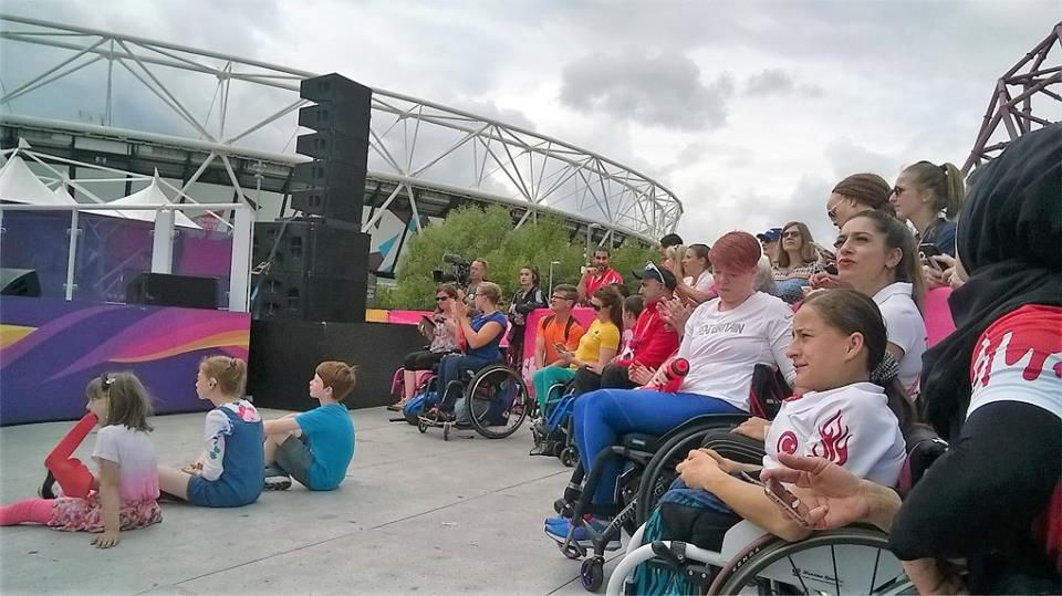 Sitting area infront of the stage in Hero's Village. Three children sitting on the ground, people sitting behind the children. People standing at the right hand side. Olympic Stadiem behinde. Dark clouds in the sky.