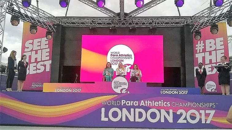 Meddle stage. Purple border along the bottem, with rainbow wave. "WORLD Para Athletics CHAMPIONSHIPS TM" written at the right. "London 2017" in bigger text under that. "SEE THE BEST" to the left of the stage, and "#BE THE NEXT" at the right. Two smartly dressed woman in suites at each side of the stage. Woman furthest right is holding a tray with meddals. Three athleets on the podium