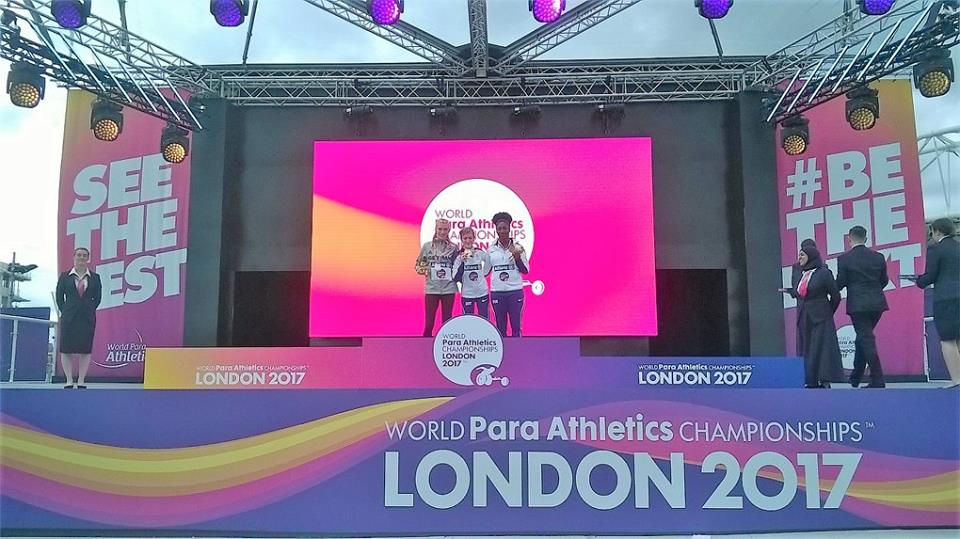 Three athleets on the podium. One woman in a suite at the left, four people in suites at the right.