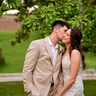 A man in a tan suit kisses a woman in a white dress on the cheek in front of a tree