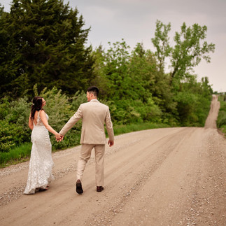 A couple walks down a gravel road line by green trees. The man is wearing a tan suit and the woman in a white dress.