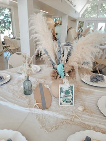 On a sand tablecloth sits surf board theme, decorations, including a tan vase with brown leafy plants coming out of it