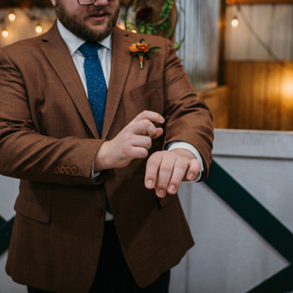 The groom stands in a brown suit, adjusting his left sleeve