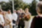 Bride and father walk down outdoor aisle, surrounded by guests. She holds a vibrant bouquet, and they both smile warmly. Sunny day vibe.