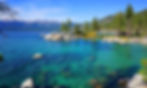 Clear blue lake with rocky shore, surrounded by pine trees and mountains under a bright sky. Relaxed, scenic atmosphere.